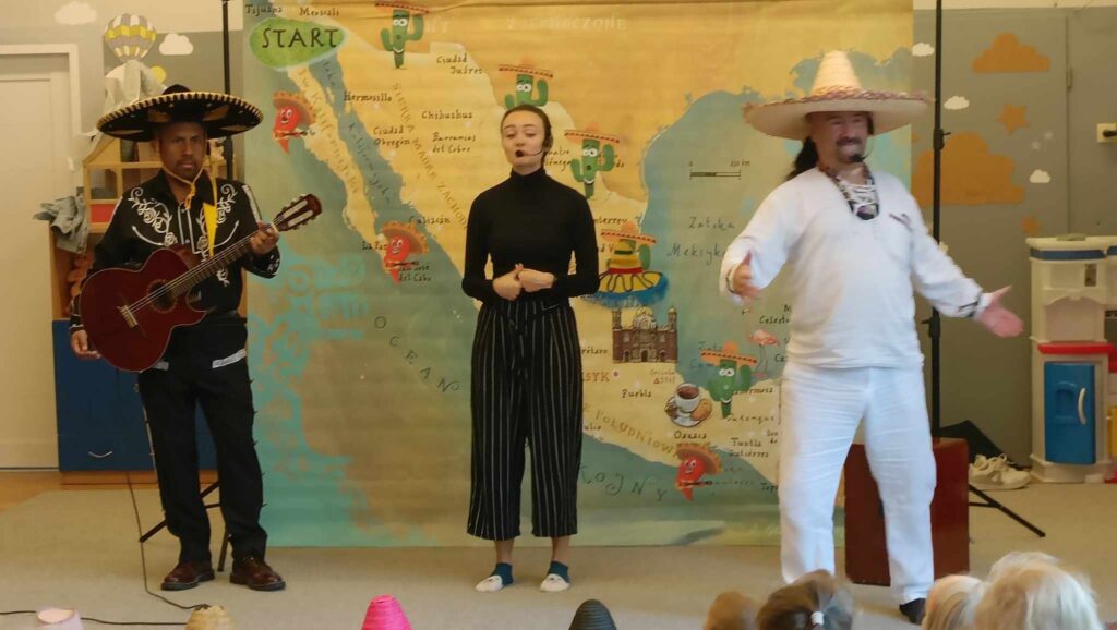 Dwóch mężczyzn w meksykańskich strojach i jedna kobieta ubrana na czarno występują dla dzieci.
